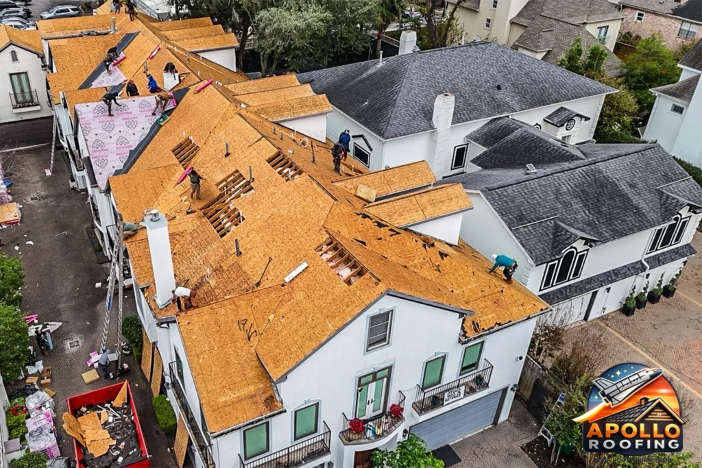 Aerial view of a residential roof under renovation, with workers removing old shingles and installing new plywood sheathing across multiple sections of the home.