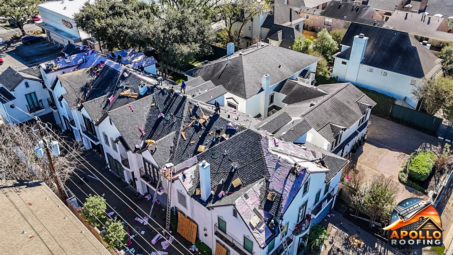 Aerial view of a residential roof under construction, with workers installing roofing underlayment and preparing multiple roof sections on a multi-level home in an urban area.