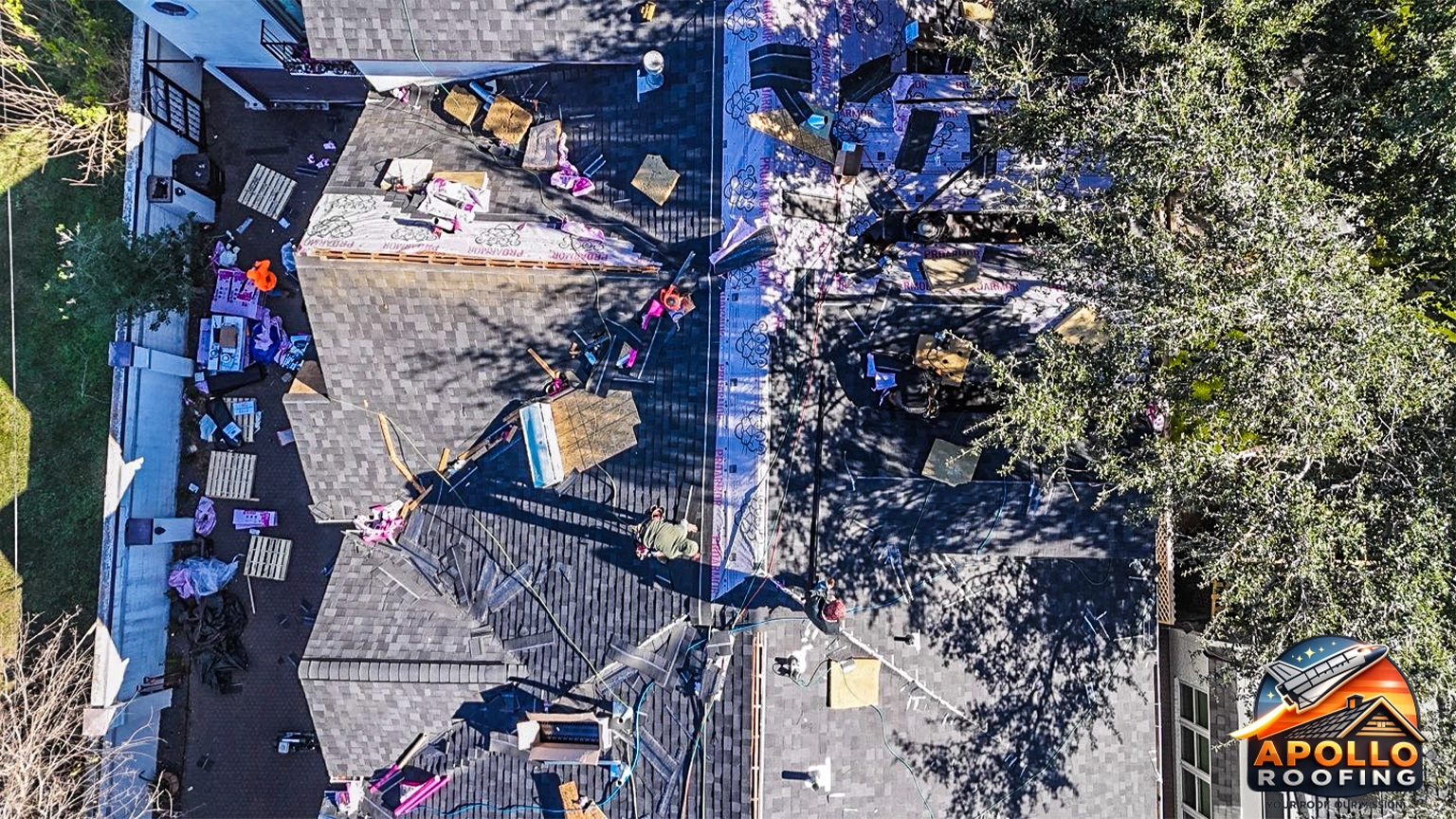 Aerial view of a residential roof under construction, with workers installing roofing underlayment and preparing multiple roof sections on a multi-level home in an urban area.