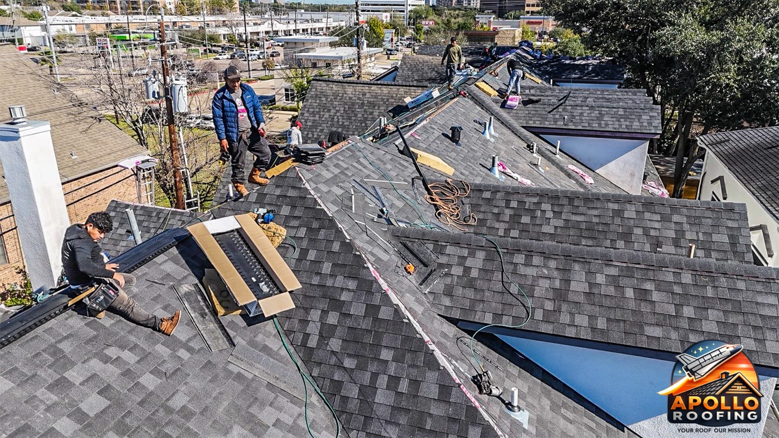 Aerial view of a residential roof under construction, with workers installing roofing underlayment and preparing multiple roof sections on a multi-level home in an urban area.