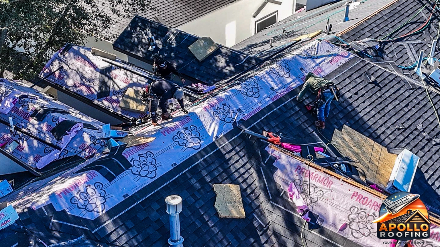 Aerial view of a residential roof under construction, with workers installing roofing underlayment and preparing multiple roof sections on a multi-level home in an urban area.