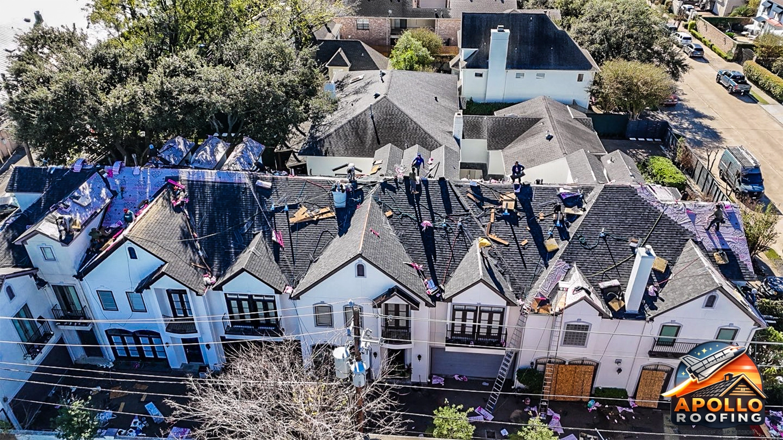 Aerial view of a residential roof under construction, with workers installing roofing underlayment and preparing multiple roof sections on a multi-level home in an urban area.