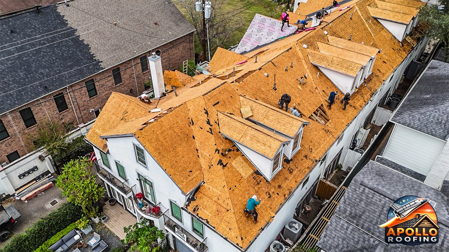 Aerial view of a large residential roof under renovation, with workers removing old shingles and installing new plywood sheathing across multiple roof sections.