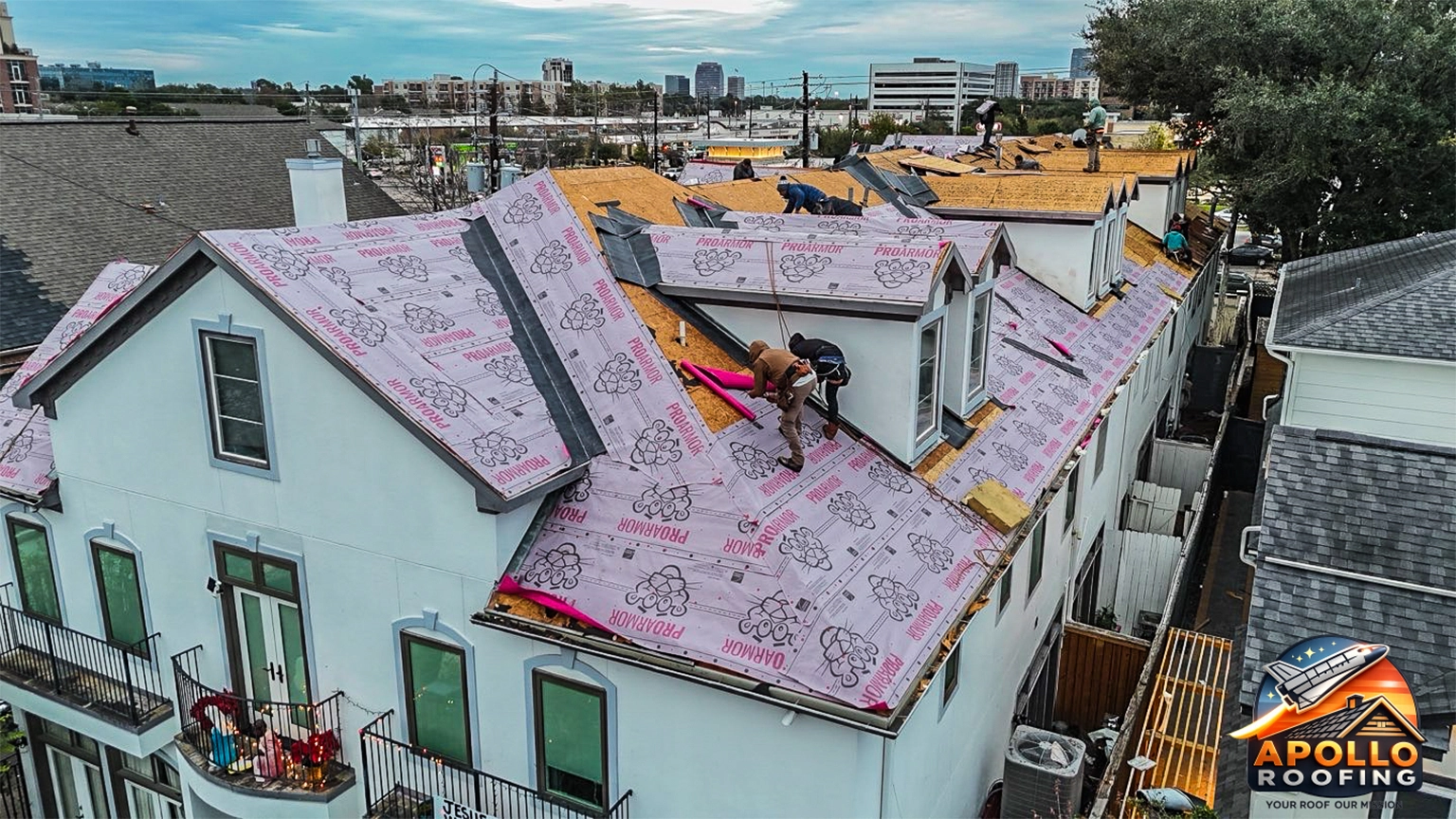 Aerial view of a residential roof under construction, with workers installing roofing underlayment and preparing multiple roof sections on a multi-level home in an urban area.