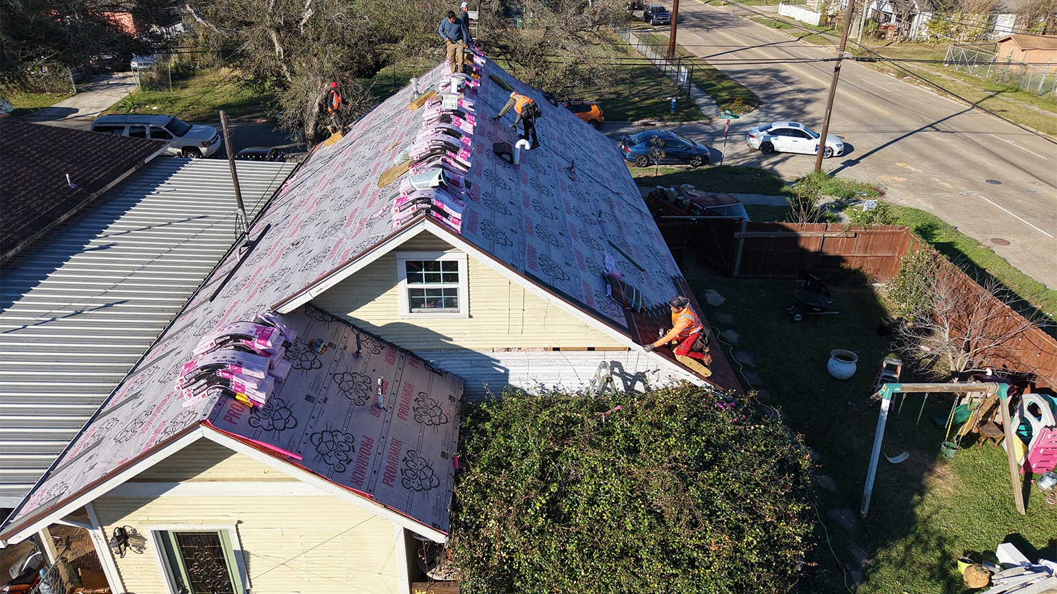 Aerial view of a small residential home with a red asphalt shingle roof, adjacent to a neighboring metal roof, surrounded by a landscaped backyard with garden beds and a walkway.