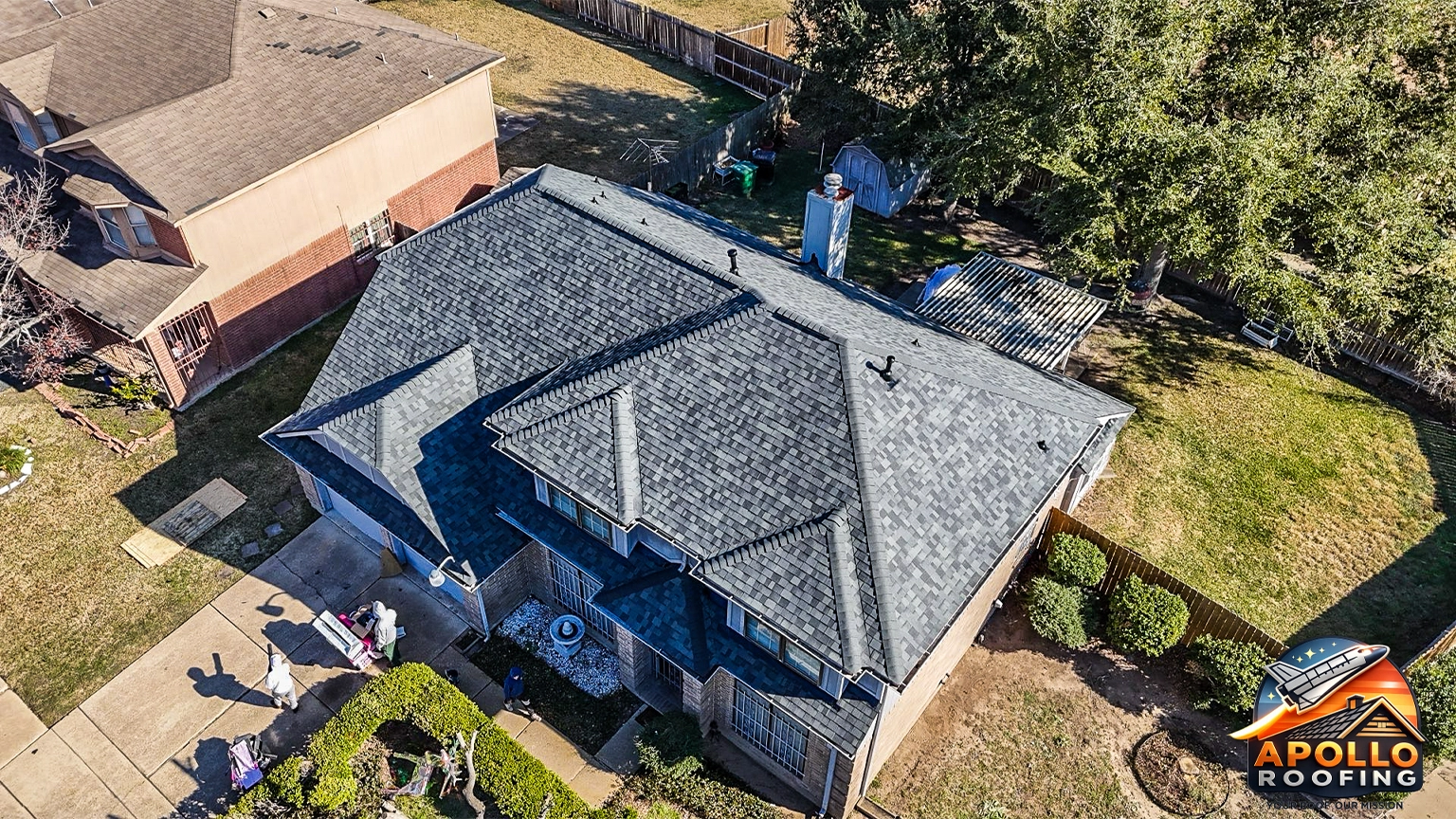 Aerial view of a residential home with a gray asphalt shingle roof, multiple slopes and dormer windows, above a brick and siding exterior.