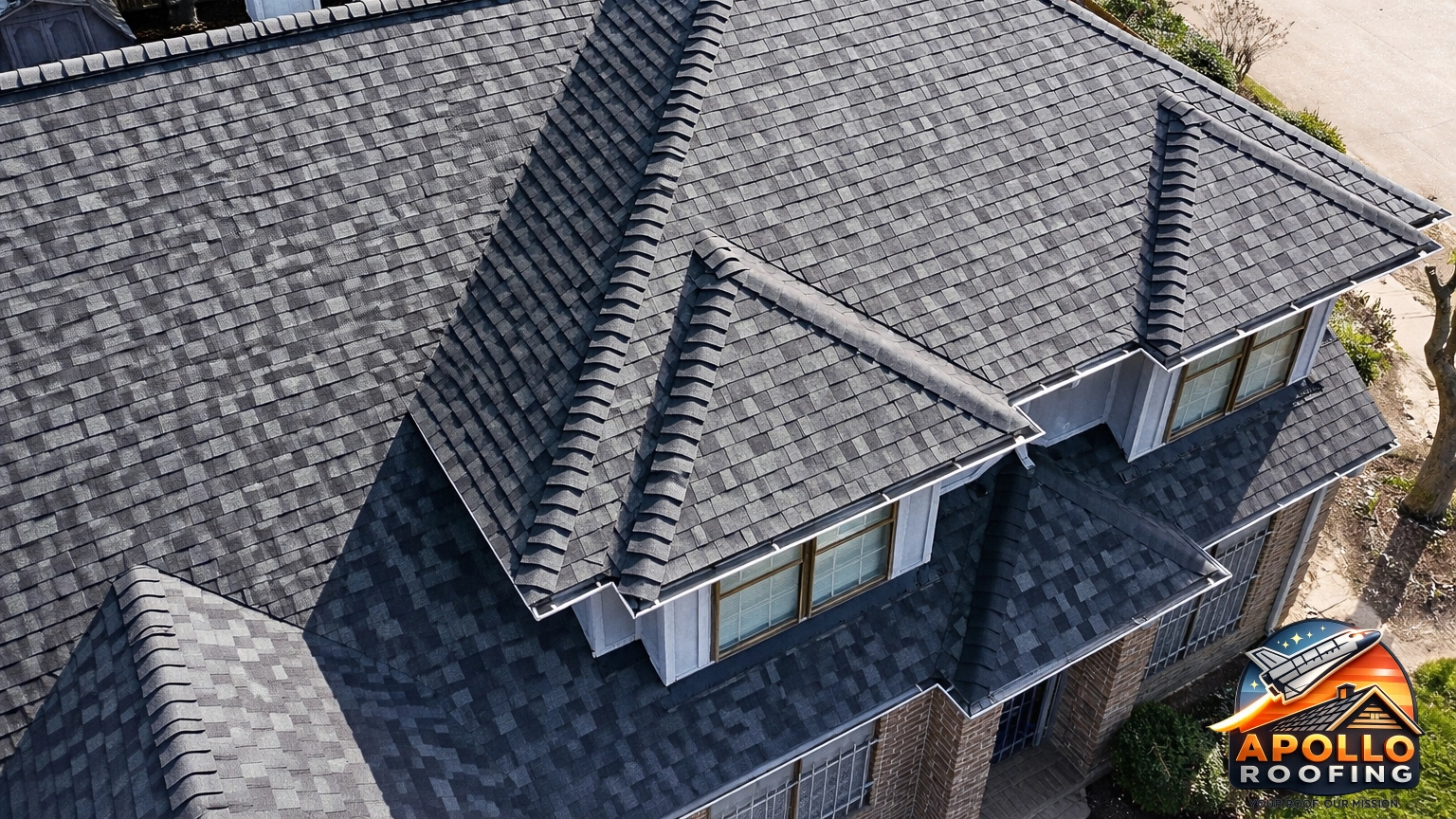 Aerial view of a residential home with a gray asphalt shingle roof, multiple slopes and dormer windows, above a brick and siding exterior.