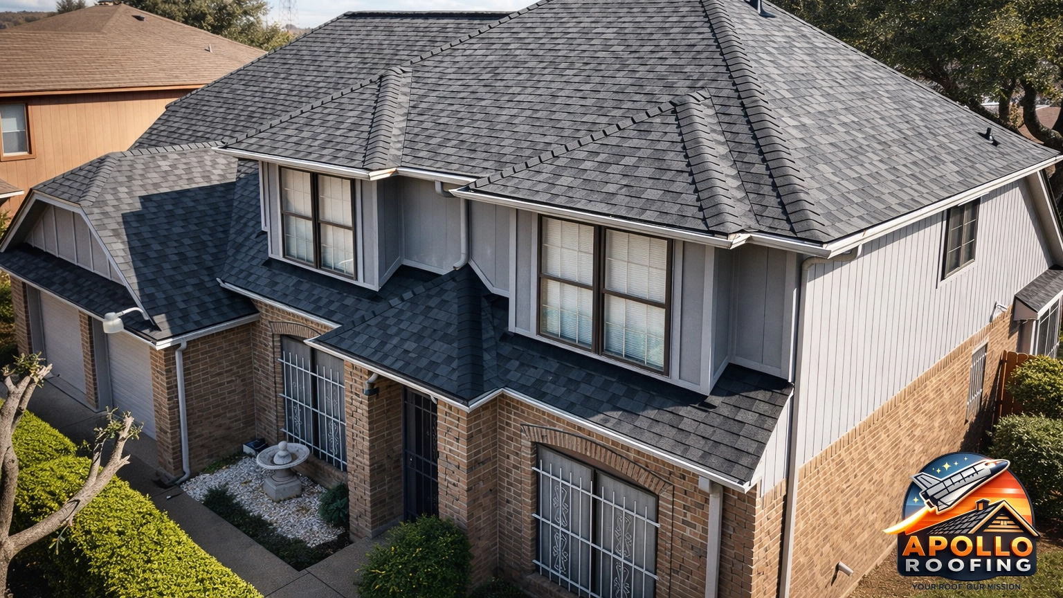 Aerial view of a residential home with a gray asphalt shingle roof, multiple slopes and dormer windows, above a brick and siding exterior.