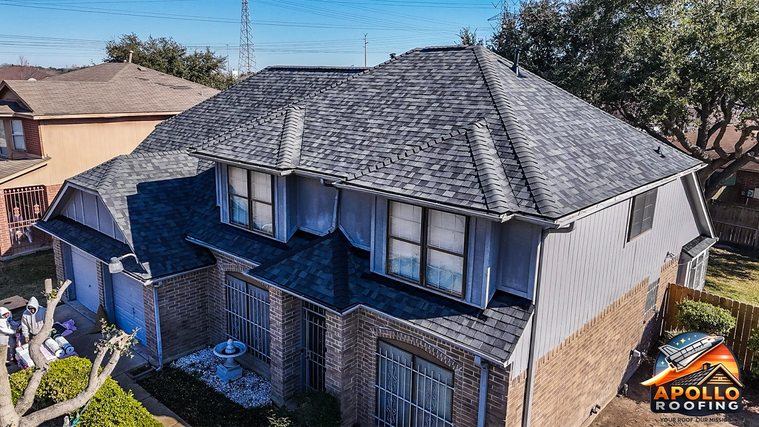 Aerial view of a residential home with a gray asphalt shingle roof, multiple slopes and dormer windows, above a brick and siding exterior.