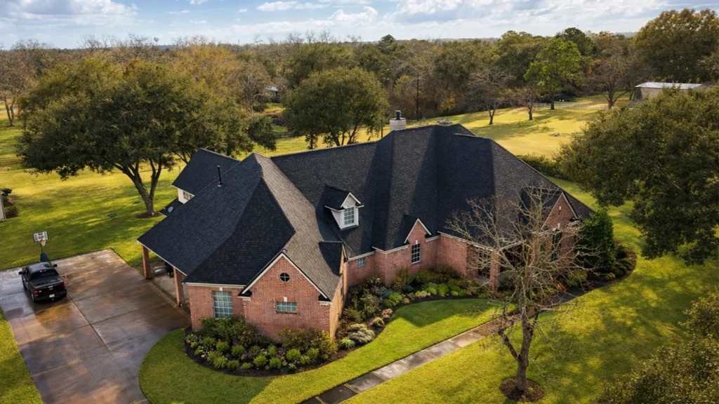 Aerial view of a large brick home with a dark asphalt shingle roof, multiple gables, and a covered driveway, surrounded by a landscaped yard and trees.