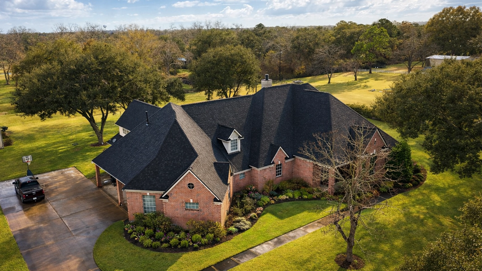 Aerial view of a large brick home with a dark asphalt shingle roof, multiple gables, and a covered driveway, surrounded by a landscaped yard and trees.