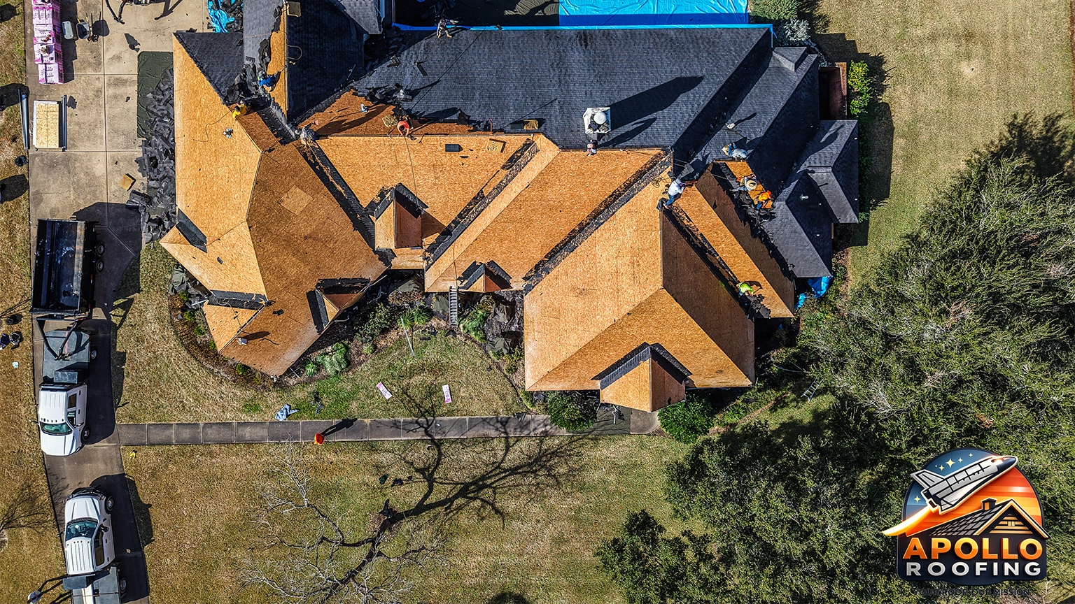 Aerial view of a large residential home undergoing roof replacement, with workers installing new sheathing and removing old shingles, debris piled near the driveway.