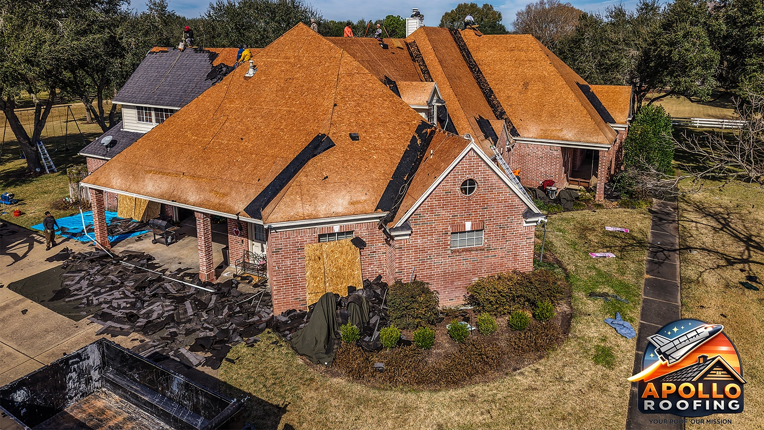 Aerial view of a large residential home undergoing roof replacement, with workers installing new sheathing and removing old shingles, debris piled near the driveway.