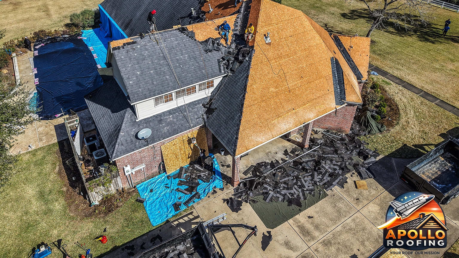 Aerial view of a large residential home undergoing roof replacement, with workers installing new sheathing and removing old shingles, debris piled near the driveway.