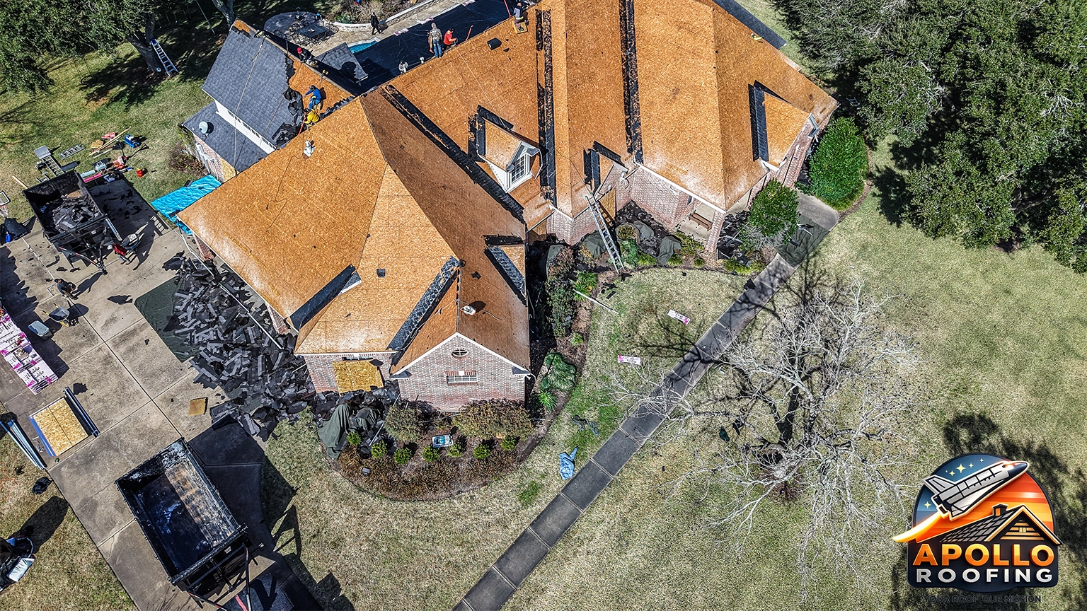Aerial view of a large residential home undergoing roof replacement, with workers installing new sheathing and removing old shingles, debris piled near the driveway.