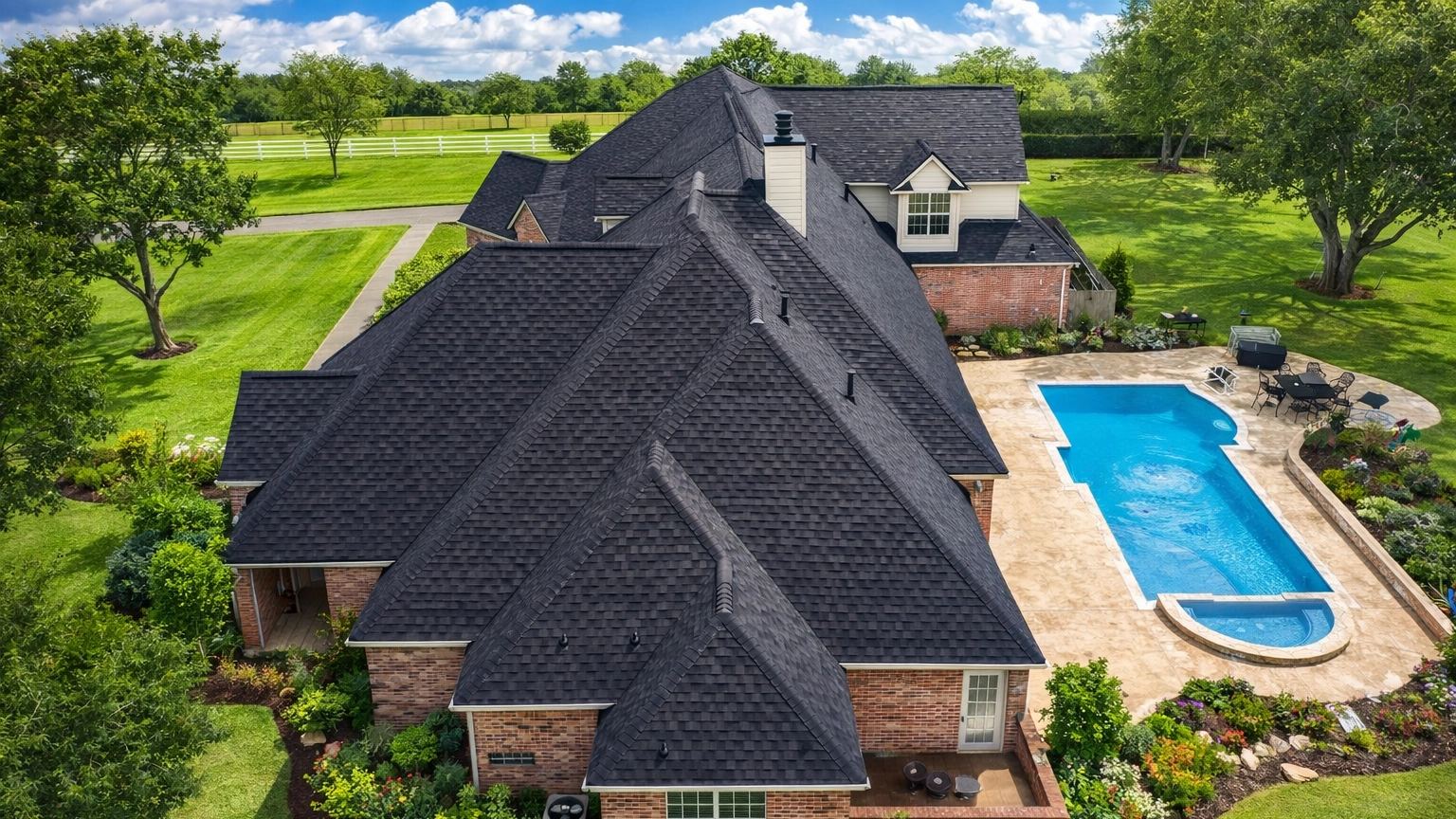 Aerial view of a large brick home with a dark asphalt shingle roof, multiple gables, and a covered driveway, surrounded by a landscaped yard and trees.