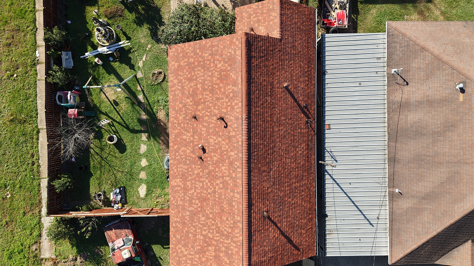 Aerial view of a small residential home with a red asphalt shingle roof, adjacent to a neighboring metal roof, surrounded by a landscaped backyard with garden beds and a walkway.