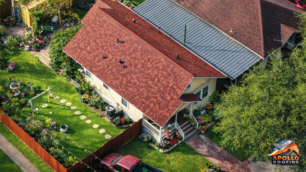 Aerial view of a small residential home with a red asphalt shingle roof, adjacent to a neighboring metal roof, surrounded by a landscaped backyard with garden beds and a walkway.